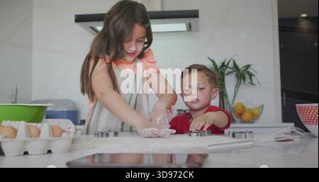 Shaping sibling pair pressing dough on kitchen counter, striped apron, red shirt, cutters, flour Stock Photo