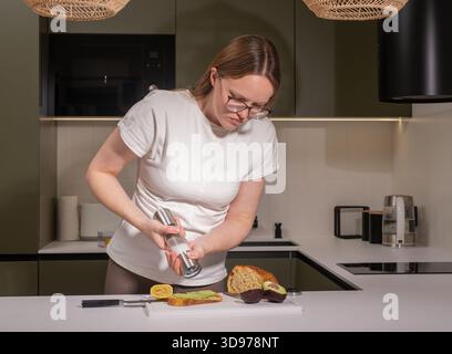 woman in a modern kitchen seasoning her avocado toast with salt and pepper, standing over a cutting board with sourdough bread, fresh avocado, lemon, Stock Photo
