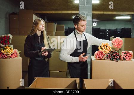 Two caucasian florists packing tulips in warehouse, filling cardboard boxes, checking colorful wrapped bouquets, using tablet for orders, warm Stock Photo