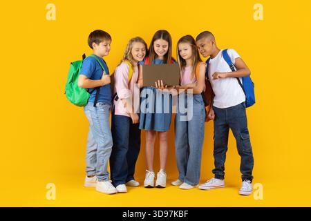Group of preteen children gathered around laptop in bright yellow studio background Stock Photo