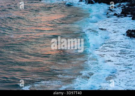 Warm sunset light reflects on the rippling ocean surface as powerful waves break against a dark, rugged rocky shore. Stock Photo
