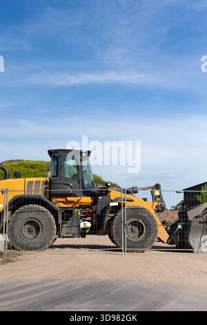 Bright yellow wheel loader stands behind metal fence on dusty construction ground. Sunlight highlights large tires, cabin and bucket while green hill Stock Photo