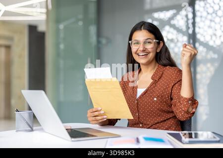 Young indian woman feeling overjoyed, holding an envelope with a letter, celebrating good news like exam results or university admission while sitting Stock Photo
