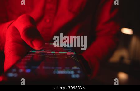 Hand holding phone while tracking stock market data and investment performance in red light Stock Photo