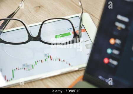 Glasses resting on a tablet displaying a financial chart and a green buy button for online trading Stock Photo