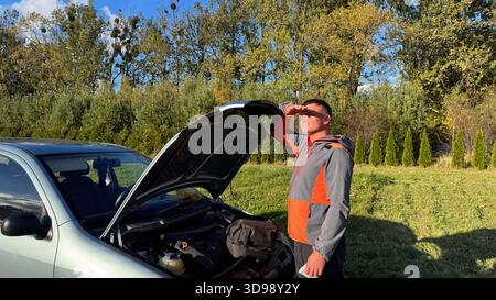 A man in a gray and orange jacket stands beside an open car hood with a tool bag inside, shielding his eyes from the sun while assessing the next step Stock Photo
