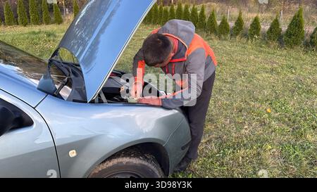 A man in a gray and orange jacket leans over the open hood of a silver car, using a tool to inspect or adjust engine components during roadside repair Stock Photo
