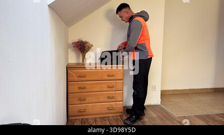 A man in a gray and orange jacket stands by a wooden dresser, zipping a black travel bag in a tidy room with wooden flooring and dried flowers in a va Stock Photo