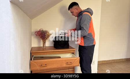 A man in a gray and orange jacket holds tools while packing a black bag on top of a wooden dresser in a simple room with an open drawer and dried flow Stock Photo