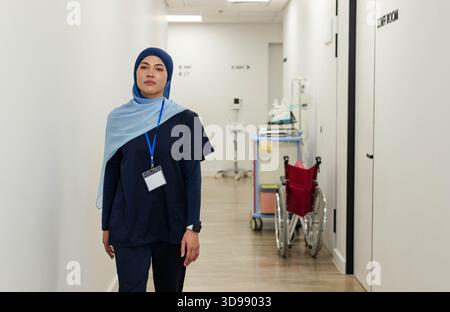 Female nurse standing in hospital corridor wearing navy scrubs, hijab, ID badge, wheelchair nearby Stock Photo