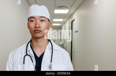 Asian male doctor standing in hospital hall wearing white coat scrubs surgical cap and stethoscope Stock Photo