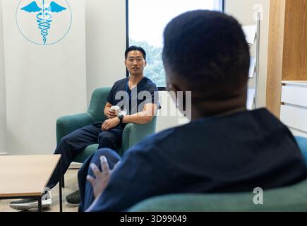 Diverse male colleagues in navy scrubs sitting in staff lounge discussing case with coffee lanyard Stock Photo