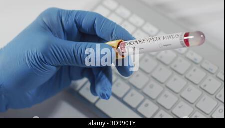 Grasping blue nitrile-gloved hand holding blood tube with yellow cap, MONKEYPOX label at lab bench Stock Photo