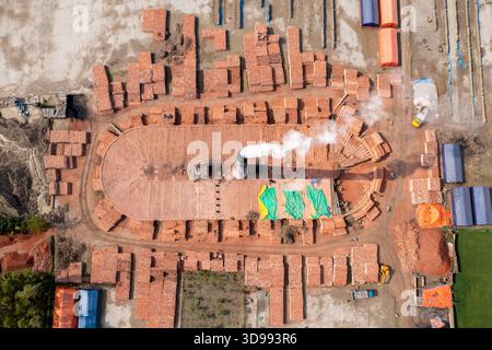 Aerial view of a brick kiln emitting smoke, surrounded by stacks of red bricks and vibrant green tarpaulins, creating a stark contrast of industry and Stock Photo