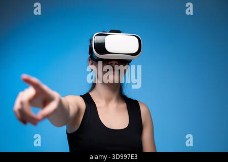 Woman wearing VR headset pointing forward while engaging with virtual interface in digital blue studio Stock Photo