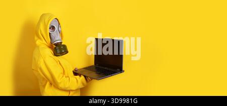 Individual in yellow hoodie and gas mask holds open laptop against vibrant yellow background. Modern, conceptual image with copy space. Stock Photo