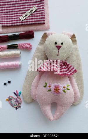 Handmade soft rabbit doll in pastel tones surrounded by pink craft supplies, delicate feminine vibe Stock Photo