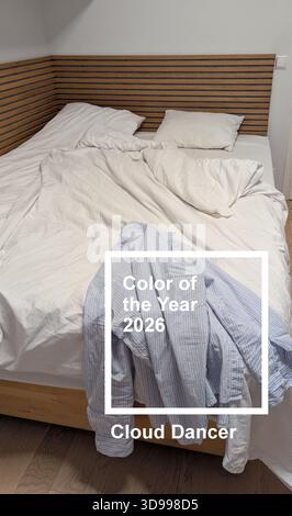 Vertical unmade bed with white bedding in cloud dancer color of the year 2026, blue striped shirt hanging on wooden bed Stock Photo