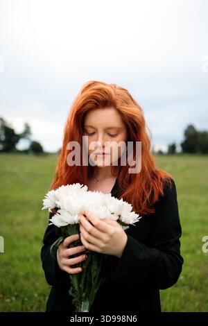 Young red haired woman holding white flowers in a quiet open field Stock Photo