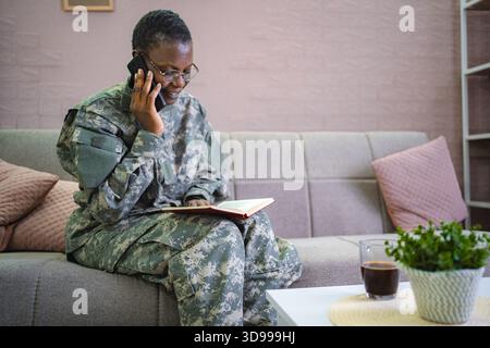 Smiling african american soldier talking on smartphone and reading book at home Stock Photo