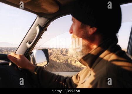 A man with a gray beard is driving a car. A dry mountain landscape is visible from the window. Sunlight falls on the driver's face Stock Photo