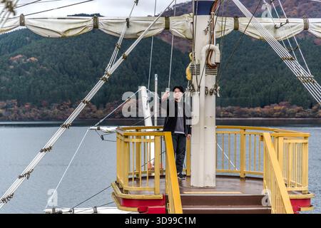 A visitor takes a photo of themselves with their smartphone on the foredeck of a sightseeing boat on Lake Ashi in Hakone, Kanagawa Prefecture, Japan. In the background are the autumnal slopes of the Hakone Mountains; Lake Ashi is a popular tourist destination from which Mount Fuji can be seen on a clear day Stock Photo