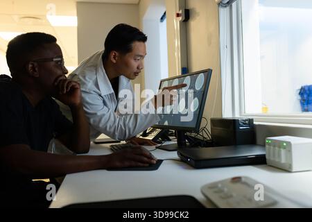 Diverse male medical staff wearing lab coat and scrubs reviewing brain scans on workstation monitor Stock Photo