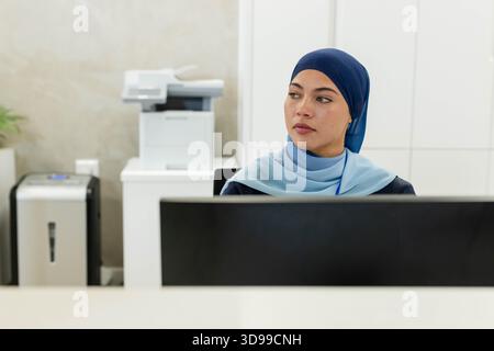 Woman wearing navy headscarf and light blue scarf sitting at reception desk using monitor Stock Photo