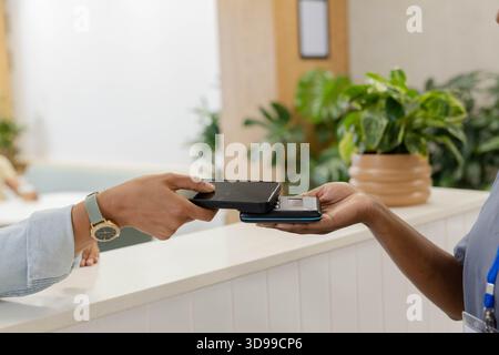 Customer and staff are tapping dark smartphone and card reader on white paneled reception counter Stock Photo