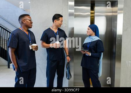 Diverse coworkers in navy scrubs talking in clinic lobby by stainless doors with coffee and tablet Stock Photo