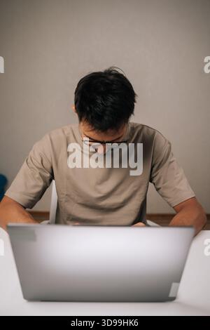 Vertical shot of focused young freelancer male wearing glasses concentrating on work on laptop, typing on keyboard in remote home office setting Stock Photo