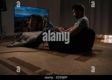 Video editor sitting on beanbag chair using laptop for editing video or photo content, with big screen tv showing footage in background. Stock Photo