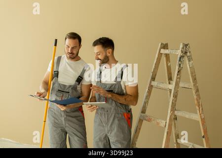 Two male workers in gray overalls are reviewing renovation plans. They are smiling and engaged in conversation, with a ladder and painting tools nearb Stock Photo