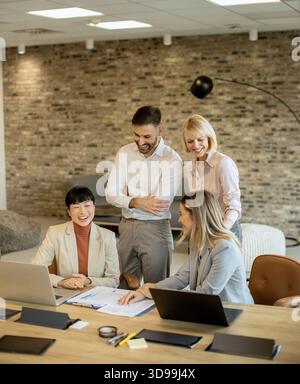 Four professionals gather in a contemporary office, sharing ideas while reviewing documents and using laptops. The atmosphere is creative and engaging Stock Photo