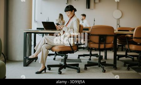 In a sleek office space, a woman in a suit checks her phone while colleagues work on laptops. The atmosphere is professional and collaborative. Stock Photo