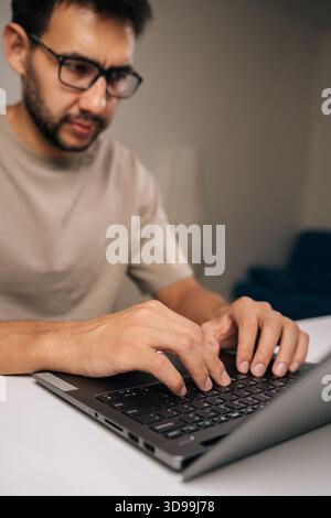 Vertical shot of focused male freelancer wearing casual clothing working remotely on laptop computer in home office workspace, representing digital Stock Photo