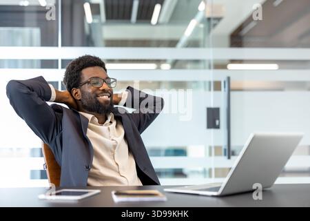 African businessman sitting comfortably at his desk with hands behind his head, smiling and feeling satisfied after completing work on his laptop in a modern office Stock Photo