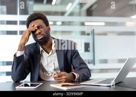 Unhappy young african american businessman experiencing headache and stress while sitting at desk in modern office, feeling overwhelmed and fatigued from professional workload Stock Photo