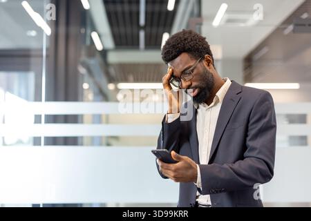 African american businessman holding a smartphone and touching his head, experiencing headache, stress, anxiety, or burnout upon receiving surprising or upsetting information in a modern office Stock Photo