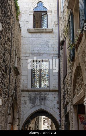 A quiet, historic alley framed by weathered stone walls. An arched window with circular glass panes overlooks a sunlit passage, evoking medieval charm Stock Photo