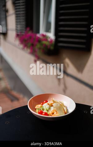 A pink bowl of shrimp salad with cherry tomatoes and crusty bread sits on an outdoor table, bathed in sunlight beside a window with a flower box, crea Stock Photo