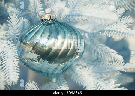 Teal glossy bauble on pastel blue Christmas tree with warm lighting. Festive decoration closeup in soft winter palette. Stock Photo