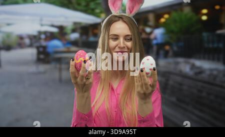 Young woman wearing bunny ears smiles outdoors while holding colorful easter eggs, embodying festive joy in a lively public setting. Stock Photo
