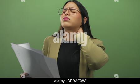 Woman appears frustrated while reviewing documents against a green background, showcasing stress as she rubs her neck, wearing glasses and a beige bla Stock Photo