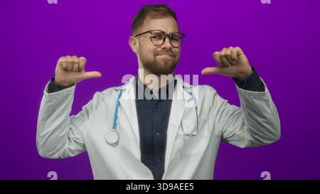 Man doctor wearing white coat with stethoscope pointing thumbs at chest in purple studio; confidence professionalism. Stock Photo