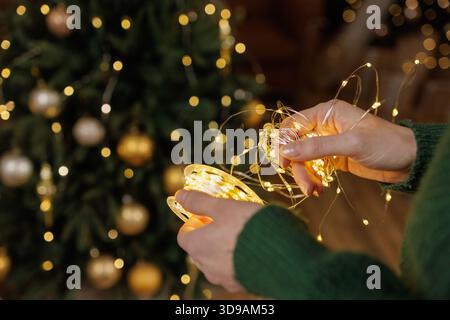 Closeup of woman in green sweater holding tangled glowing string lights with decorated Christmas tree and golden bokeh behind Stock Photo