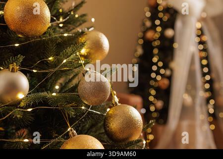 Close view of decorated Christmas tree with golden baubles and string lights and blurred holiday tree in cozy home interior Stock Photo