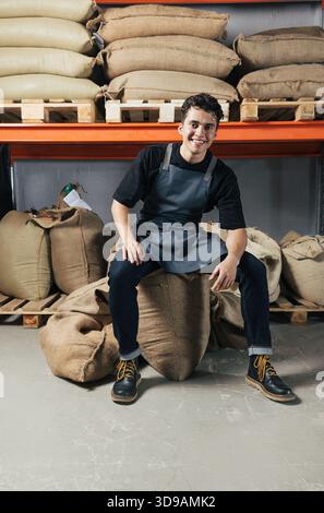 Exhausted warehouse worker sitting on a coffee bag and looking at camera. Young worker in apron sitting in warehouse. Stock Photo