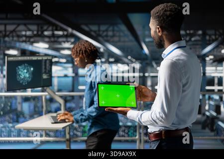 African american engineer uses chroma key display on tablet in data center, analyzing performance outputs standing on industrial platform. Server hub engineering AI configurations. Stock Photo