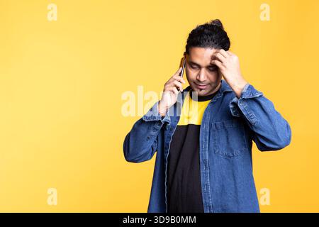 A friendly person with man bun calling his friend on phone call to gossip, sharing information in a remote conversation against studio background. Indian cheerful man with a modern lifestyle. Stock Photo